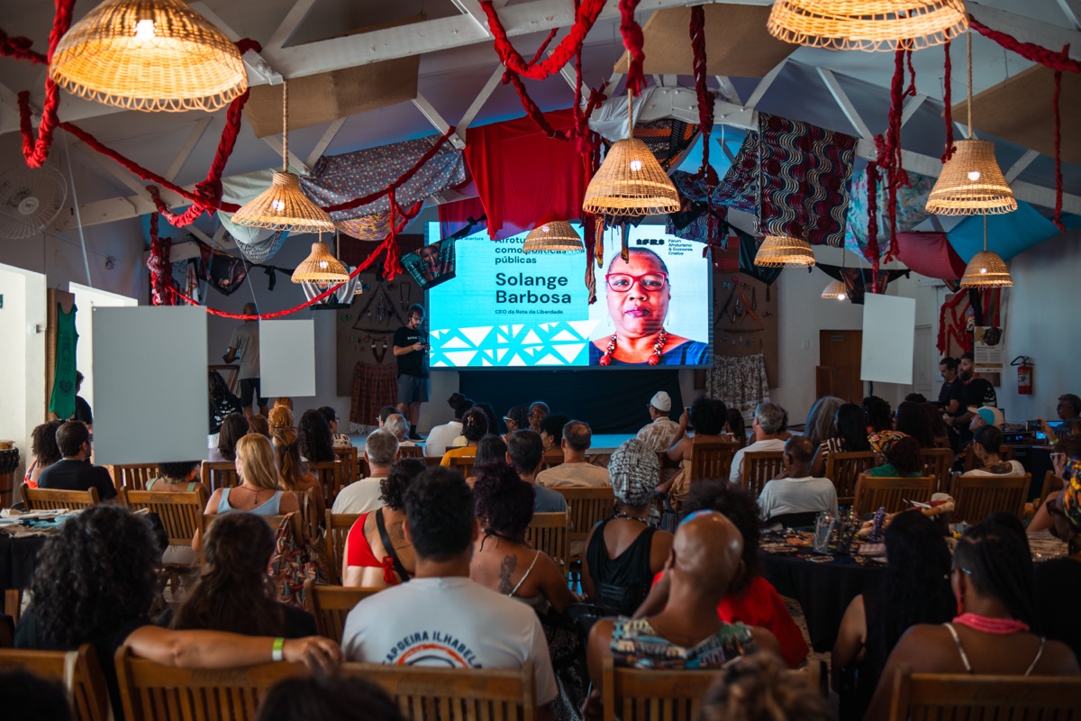 Solange Barbosa, palestrante, projetada em tela; plateia diversa sob decoração caiçara-afro